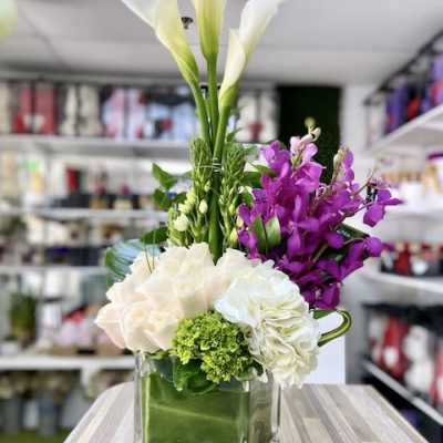 Tall white calla lilies with purple orchids and pale roses in a green-lined glass cube vase.