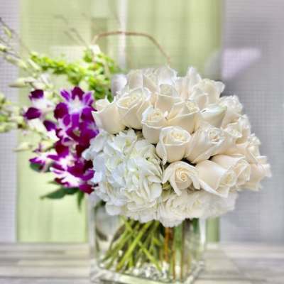 White roses and hydrangeas in a clear cube vase with purple orchids and green Bells of Ireland in the background.