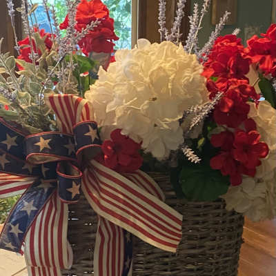 Red and white floral arrangement in a woven basket with an American flag ribbon