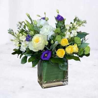 Mixed bouquet of white, yellow, and purple flowers in a square glass vase