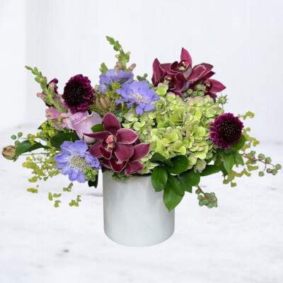 Mixed purple and green floral arrangement in a white cylindrical vase