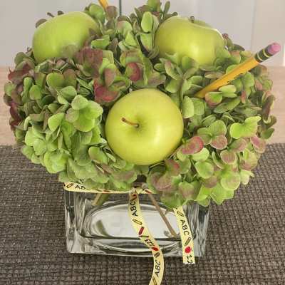Green hydrangea arrangement with apples and pencils in a glass vase