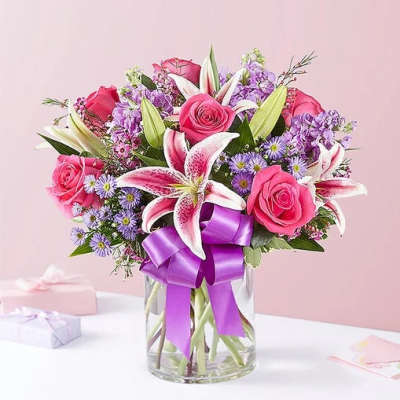 Pink roses and lilies in a glass vase with a purple ribbon