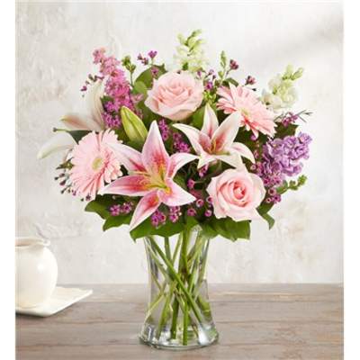 Pink lilies, roses, and gerbera daisies with lavender blooms arranged in a clear glass vase