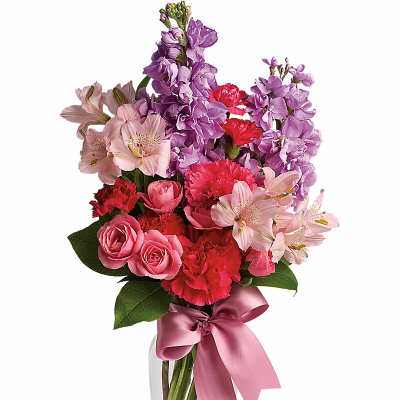 Tall mixed bouquet of pink roses, red carnations, and purple stock in a clear vase with a pink ribbon.