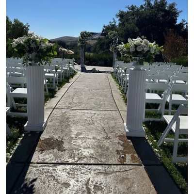 Outdoor wedding aisle with white flower arrangements on tall white columns and an arch in the distance