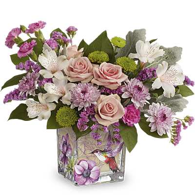Short arrangement of pink roses, white lilies, and purple blooms in a glass cube vase with hummingbird art.