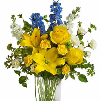 Tall glass vase of yellow lilies and roses with blue delphinium and white blooms