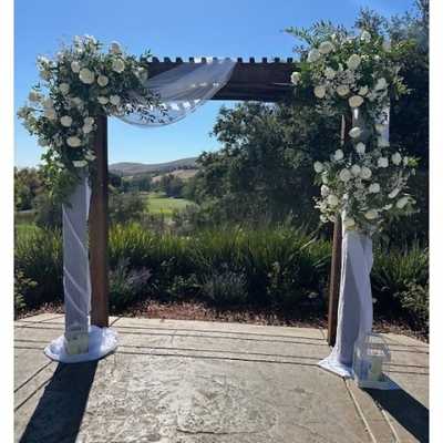 Outdoor wooden arch with white floral arrangements and sheer white draping fabric