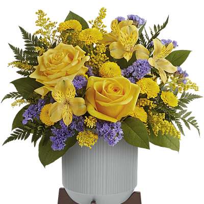 Arrangement of yellow roses with mixed yellow and purple flowers in a gray pot on a wooden stand.