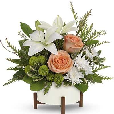 Low arrangement of white lilies, peach roses, white mums, and green button flowers in a white bowl on a wooden stand.