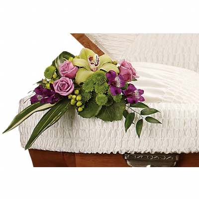 Small purple, green, and orchid floral arrangement resting on the edge of a white cushioned casket