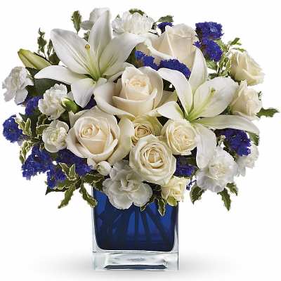 Compact arrangement of white lilies, roses, and carnations with blue accents in a blue glass cube vase