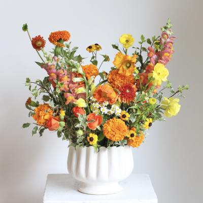 Bright mixed bouquet in a white pedestal vase with orange, yellow, and pink blooms.