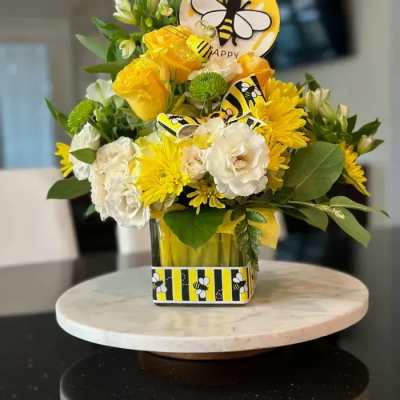 Yellow and white flower arrangement with bee-themed accents in a glass cube vase on a marble stand