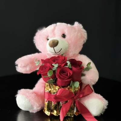 Pink teddy bear holding red roses and chocolates with a ribbon