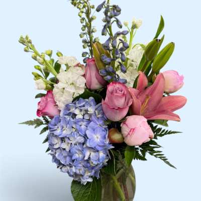Mixed bouquet of pink roses, blue hydrangea, white blooms, and lilies in a glass vase