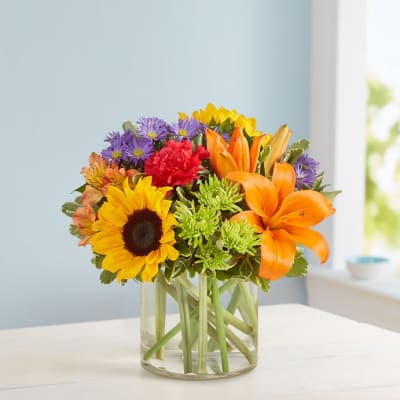 Mixed bouquet with sunflowers, lilies, and purple daisies in a glass vase