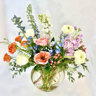 Mixed bouquet in a round glass vase with roses, ranunculus, and tall blue and white spikes.