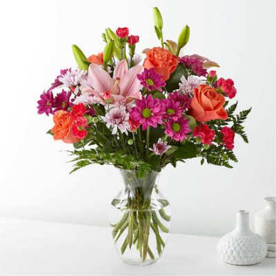Mixed bouquet of pink lilies, orange roses, and bright daisies in a clear glass vase