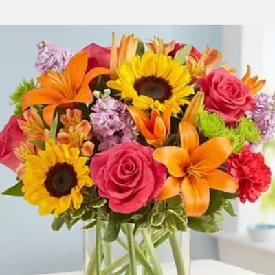Bright mixed bouquet in a clear glass vase with sunflowers, roses, lilies, and carnations.