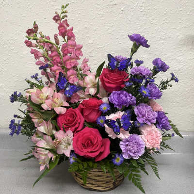 Basket arrangement of pink roses, purple carnations, and pink snapdragons with butterfly picks