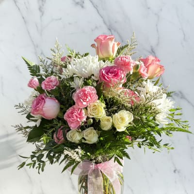 Pink and white bouquet of roses, carnations, and daisies in a glass vase with a ribbon.
