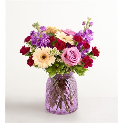 Mixed bouquet of pink, purple, and cream flowers in a purple glass vase