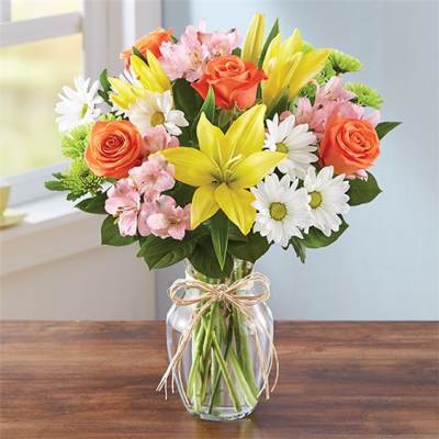 Bouquet of yellow lilies, orange roses, pink flowers, and white daisies in a glass vase
