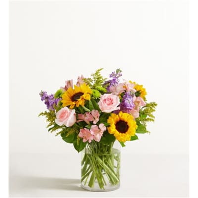 Mixed bouquet of sunflowers, pink roses, and purple blooms in a clear glass vase