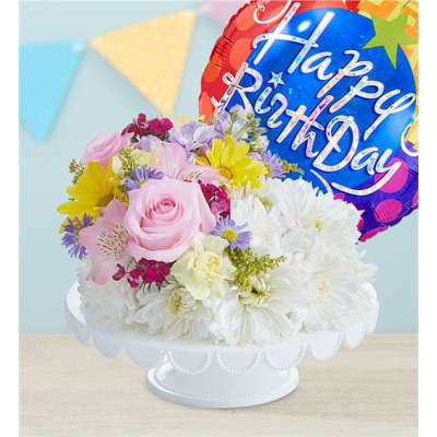 Mixed pastel flower arrangement with a birthday balloon in a white pedestal bowl