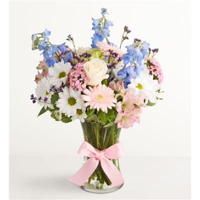 Mixed bouquet of blue, white, and pink flowers in a glass vase with a pink ribbon