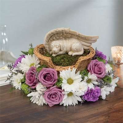 Purple roses and white daisies surround a wicker basket with a sleeping angel dog figurine.