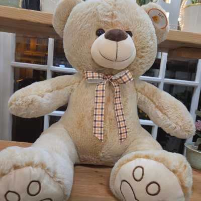 Large beige teddy bear with a plaid bow sitting on a wooden shelf