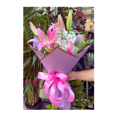 Hand-tied bouquet of pink lilies, white flowers, and pink roses wrapped in purple paper with a large bow.