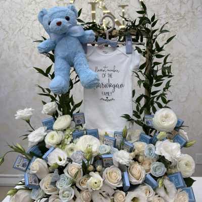 White floral baby gift arrangement with a blue teddy bear and baby onesie