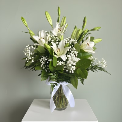 White lilies arranged in a glass vase with baby's breath and a white ribbon
