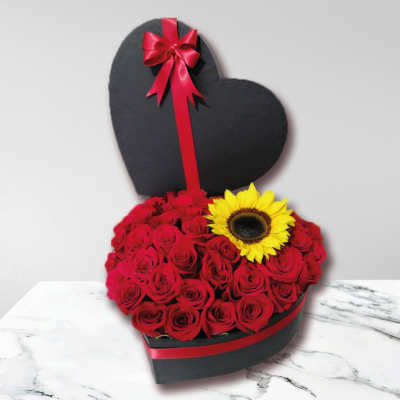 Heart-shaped black box filled with red roses and a single yellow sunflower accent.