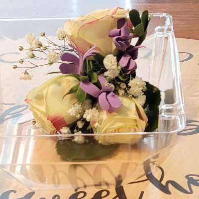 Small bouquet of pale yellow roses with purple accents in a clear plastic box