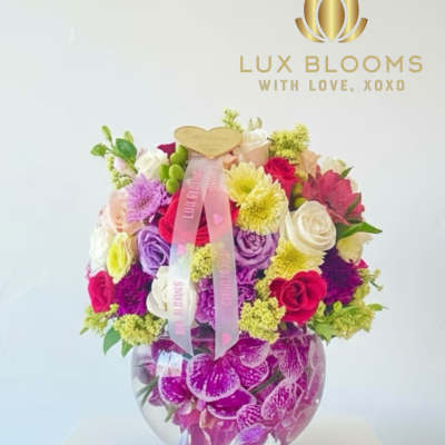 Colorful mixed flower arrangement in a round glass vase with a pink ribbon
