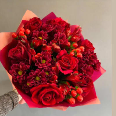 Handheld bouquet of red roses and burgundy flowers wrapped in pink paper