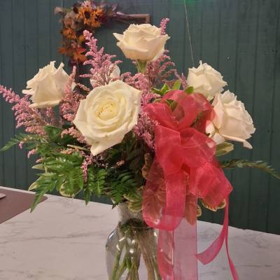 White roses and pink filler flowers in a clear glass vase with a pink ribbon