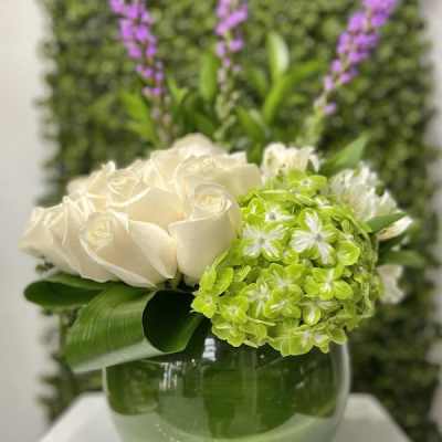 Low arrangement of white roses and green hydrangea in a round green glass vase with tall purple flower spikes.
