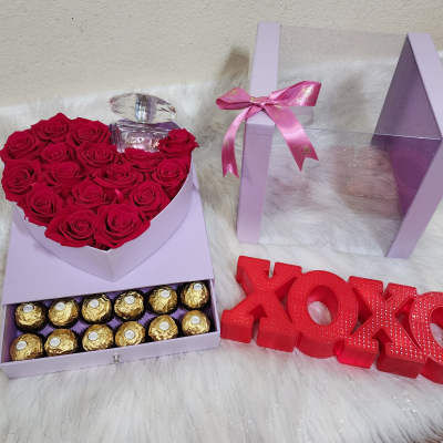 Heart-shaped box of red roses with a perfume bottle and chocolates beside XOXO decor