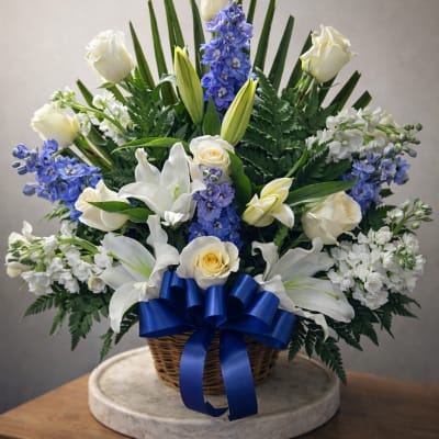 White roses and lilies with blue flowers in a wicker basket tied with a blue ribbon.