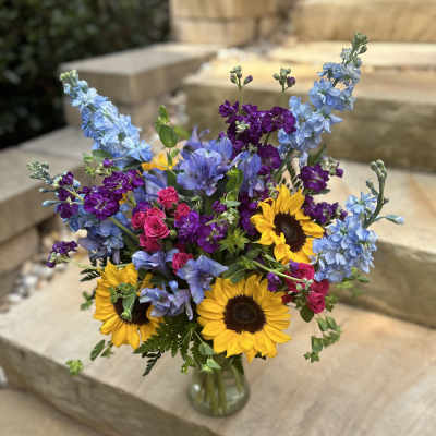Bouquet of sunflowers, blue delphinium, purple blooms, and pink roses in a glass vase