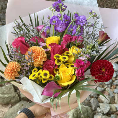 Handheld bouquet of multicolored roses, dahlias, and purple blooms wrapped in pink paper