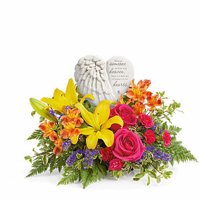 Low arrangement of yellow lilies, bright pink roses, and mixed blooms with a white angel wing heart keepsake.