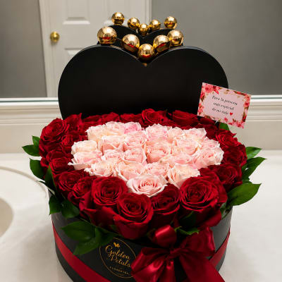Heart-shaped box of red and blush roses with a red ribbon and gold topper
