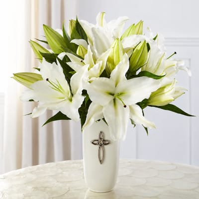 White lilies arranged in a tall white vase with a cross emblem